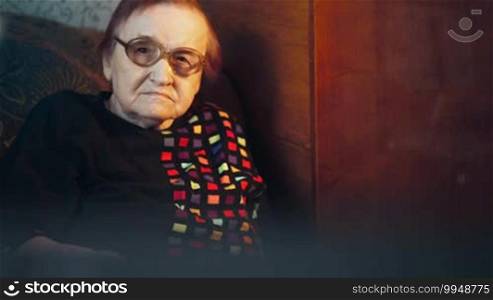 Elderly woman in glasses sitting on the sofa watching TV and changing channels with remote control