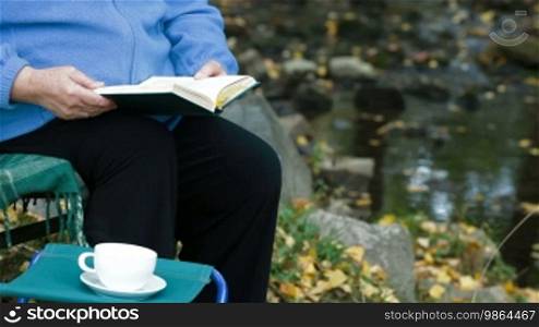 Elderly woman reading a book with a cup of coffee in the park. Unrecognizable person, side view