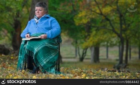 Elderly woman sitting in a chair, sheltered by a blanket and reading a book, front view