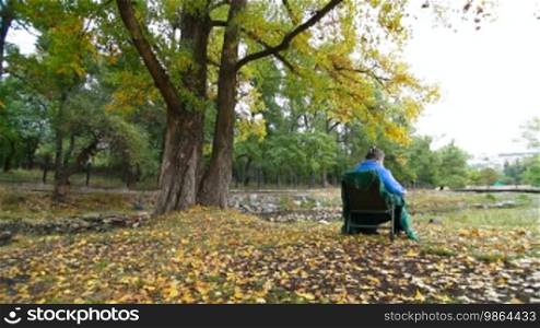 Elderly woman sitting on a chair at autumn park. Tripod, Wide Shot, Rear View