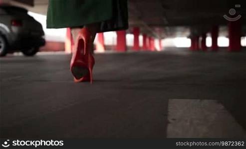 Elegant female legs in orange high heel shoes walking to parked car in covered parking garage. Woman in green coat and stylish shoes approaching parked auto in parking lot and pressing remote control car alarm system to open door. Slow motion.