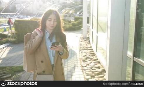 Elegant young Asian woman carrying shopping bags over her shoulder and surfing the net with a mobile phone in the city street after shopping at sunset. Slow motion. Cheerful brunette female using a smartphone app to shop online while walking outdoors.