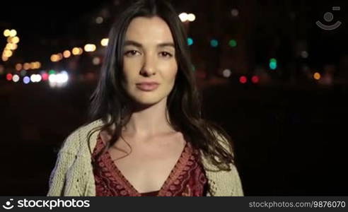 Elegant young brunette with deep brown eyes posing and gazing at camera with mysterious look on night city street. Portrait of sensual woman with long hair playing with cam and looking with shy smile seductively at night. Slow motion. Stabilized shot.