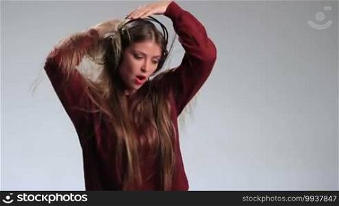 Emotional hipster girl in headphones enjoying her favorite music and dancing on white. Excited attractive woman in positive playful emotion listening to music in headphones, playing with her amazing long hair, swinging to the beat, and smiling cheerfully.