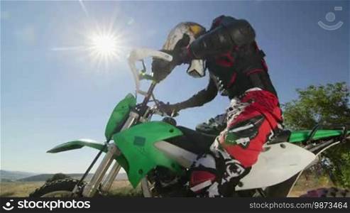 Enduro racer starting engine of his dirt bike riding away against sun side view