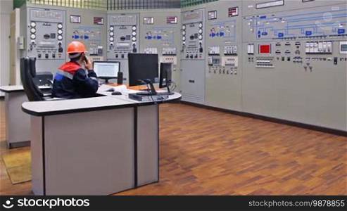 Engineer sits and talks on phone near main control panel of gas compressor station, then looks at monitor while making notes in registry