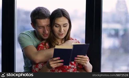 Enjoying every moment together. Closeup affectionate young couple reading a book together with happy facial expressions. Cheerful man embracing stunning brunette female gently from behind while spending leisure at home.