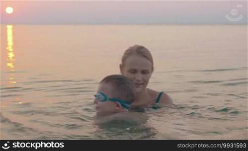 Evening with mom in the sea. Boy learning to swim with mother's help. He is doing great and happy woman showing thumbs up.