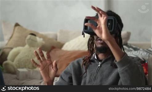Excited African American man with dreadlocks sitting on the floor and using VR headset glasses. Hipster in virtual reality headset surfing the net and gesturing. People using new trends technology.