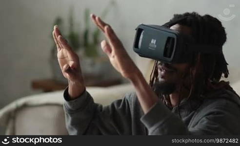 Excited African American man with dreadlocks wearing virtual reality headset glasses, playing video games in a modern design apartment close-up. Headset VR device playing with smartphone. Slow motion.