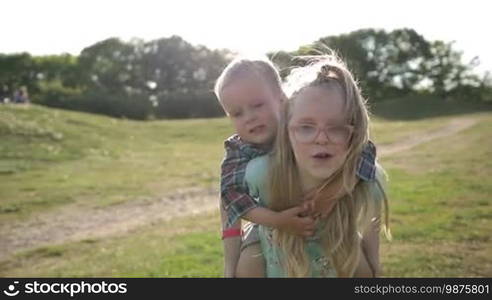 Excited sister giving joyful toddler brother piggyback ride on park lawn on sunny day. Cute young girl carrying her little brother on back and running while siblings playing, smiling, laughing and having fun outdoors. Slow motion. Stabilized shot