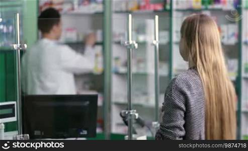 Experienced pharmacist in white coat counseling female customer in modern pharmacy, view of the pharmacist over the client's shoulder. Handsome friendly drugist with stethoscope selling medicine at the drugstore. Health care and pharmacology concept