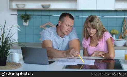 Family budget and finances. Serious couple reviewing their bank accounts using a laptop computer and documents in a modern kitchen. Woman and man doing paperwork together, paying taxes online on a notebook PC.