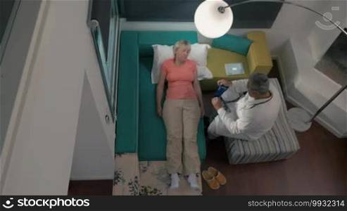 Family doctor came to the home of her mature patient to check up. She is lying on the couch, he is recording the case on a tablet.