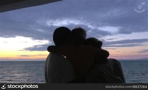 Family hugs: Son is hugging mother and father against sea sunset. Father is holding son on his hand. View from back.