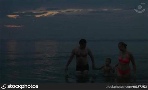 Family is swimming in the sea and holding hands after sunset on a dusky sky background. Mother, father, and son are leaving the water. Location: Piraeus, Greece