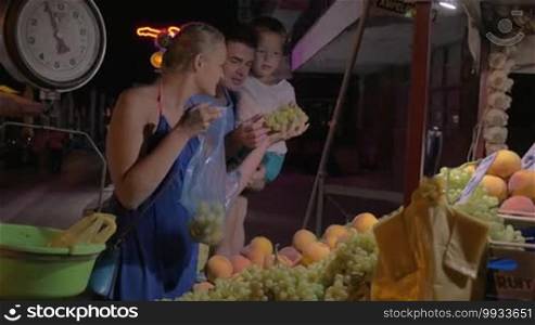 Family of three shopping for fruit on the market in late evening. Mother taking grapes and peaches, father holding son in arms. Seller weighing the fruit