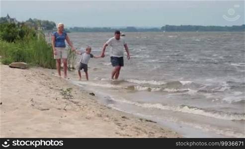 Family with child spending time together on beach holidays. Mother, father and son holding hands and walking along the beach shore leaving footprints on the sand during summer vacation