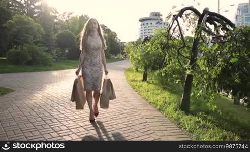 Fashionable blonde woman in red high heels and bodycon midi dress with shopping bags walking along city street. Beautiful sexy female carrying shopping bags after day shopping in the rays of setting sun. Slow motion. Steadicam stabilized shot.