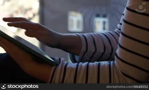 Fast dolly shot of a woman working with a tablet PC sitting by the window on the high floor. Defocused city view in the background