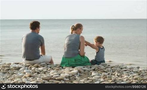 Father, mother, and son sitting on pebble beach by the sea. Woman and child playing, man enjoying the view, boy showing something in water