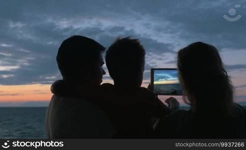 Father stands with son in his arms, and mother is photographed on tablet of sunset and sea in city Perea, Greece. Then mom laughed and kisses his son