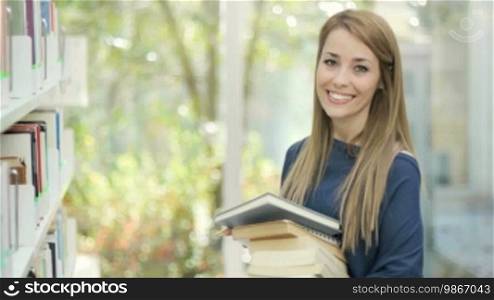 Female blonde college student taking book from shelf in library and looking at camera