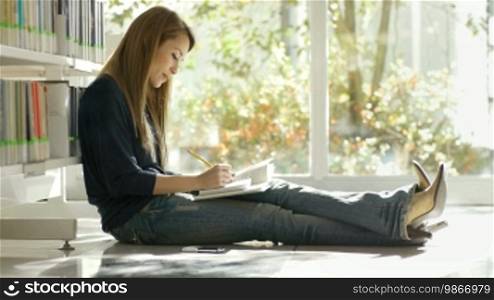 Female college student sitting on floor in library, reading book and taking notes. Copy space