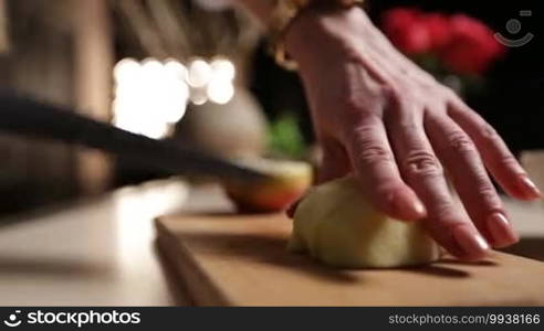 Female hand with knife slicing into pieces peeled apple on wooden chopping board in kitchen while preparing healthy fruit smoothie. Middle section of woman chopping apple on cutting board. Side view closeup.