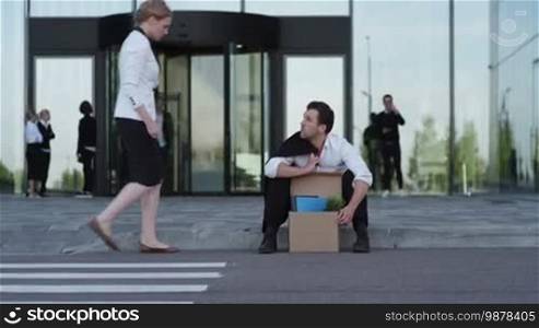 Fired business man sitting on the street near office building with box of his belongings. He lost work. Business woman comforts and encourages him