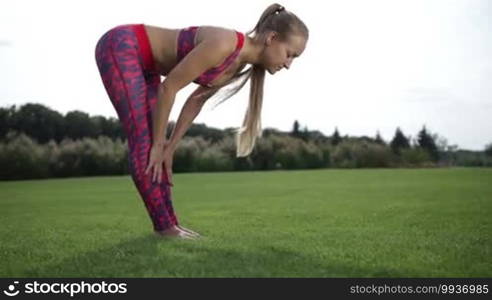 Fit young woman practicing stretching exercise, doing standing head to knees posture, uttanasana pose outdoors in the park on summer day