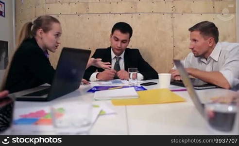 Five young business people discussing a new project using a laptop and touchpad to note ideas during a meeting at the office. A young beautiful woman is gesturing and discussing while her coworkers listen to her sitting at the office table.
