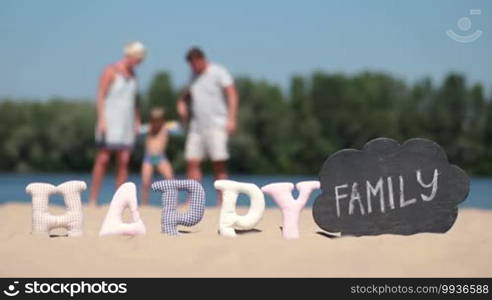 Foreground word happy made of fabric padded letters and word family written with chalk on a black board. Blurred background of family with kid playing on the beach during summer vacation