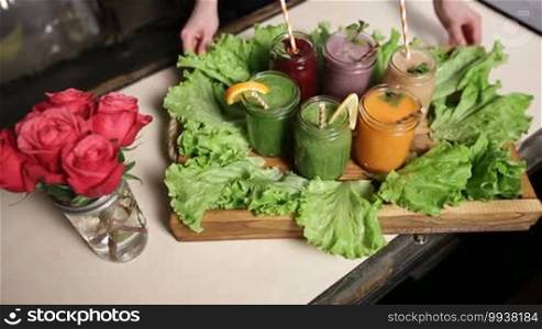 Freshly blended smoothies of various colors and tastes in jars served on a wooden tray decorated with green salad. Top view. Woman serving a rustic wooden tray with an assortment of fruit and vegetable smoothies in mason jars. Healthy eating and dieting.