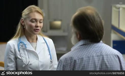 Friendly attractive female physician talking to ill stressed senior male patient and giving treatment advice in clinic. Cheerful woman doctor encouraging elderly man and touching patient's shoulder to show support and empathy after medical exam.