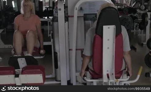 Front and back view of two senior women working out on back extension and inner thighs fitness machines in the gym