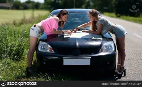 Front view of cheerful women on high heels on vacation. Car parked on a roadside in the countryside and females bent over looking at map on the car hood for directions of road trip