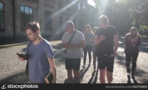 Generation Z. Group of hipster college friends browsing the net with mobile phones while walking together on university campus. Students busy with smartphones on the way to college. Slow motion. Steadicam stabilized shot.