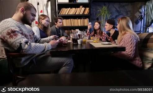 Generation Z. Group of young teenage friends sitting at a wooden table at a coffee shop using smartphones and chatting. Hipster teenagers using modern gadgets, browsing the internet and social media with mobile phones, scrolling webpages, and texting messages.