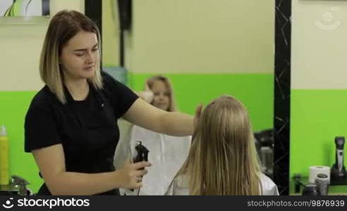 Good looking little girl getting her hair wet before getting a haircut in beauty salon. Beautiful female barber spraying water on client's hair in hairdressing salon.