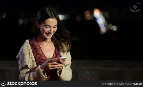 Gorgeous young smiling brunette woman texting message on smartphone at night over colorful city lights bokeh background. Elegant long brown hair female typing on mobile phone while relaxing outdoors in city at night.