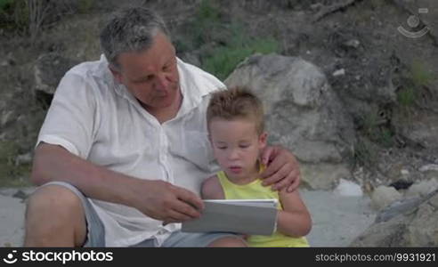 Grandfather and little grandchild spending time outdoors. Boy using tablet computer while grandpa embracing and talking to him