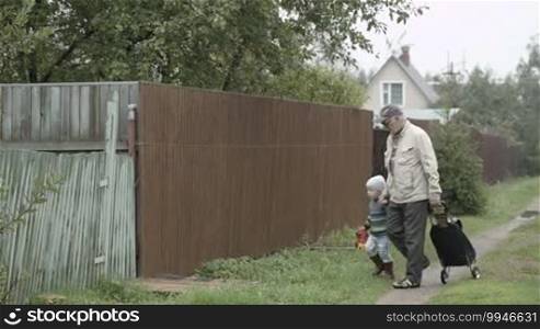 Grandpa and his little grandson coming into house gate in the village. Old man rolling a trolley bag.