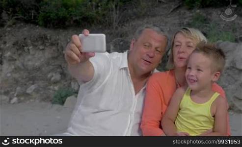 Grandparents are sitting outdoors with their grandson. They are taking a selfie shot with a smartphone.