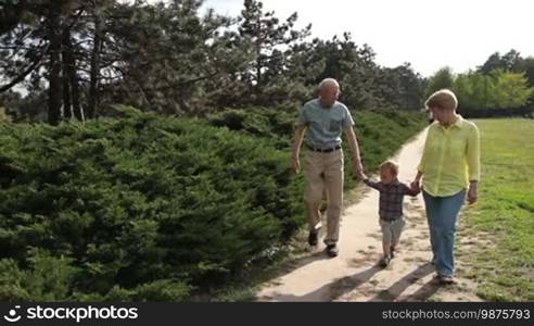 Grandparents enjoying their retirement: Senior woman and old man spending time with their grandson in the park. The old people holding boy's hand and taking a walk on a sunny day outdoors. Slow motion. Steadicam stabilized shot.