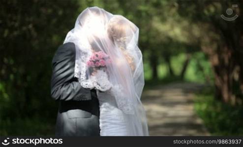 Groom kisses the bride, having covered under a veil, on a park alley