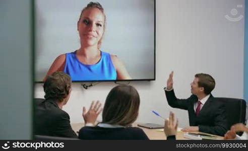 Group of business people meeting in corporate conference room, doing a video conference call with a female colleague on a big TV monitor. Wide shot