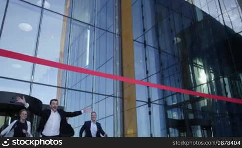 Group of happy business people running from office building crossing red ribbon finish line