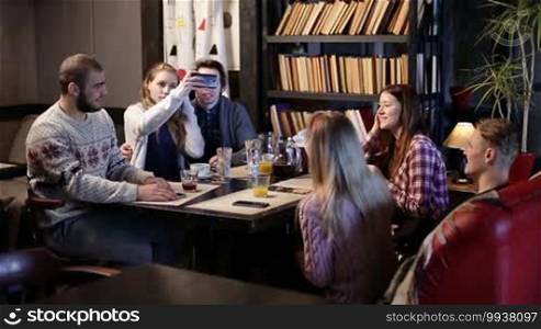 Group of smiling hipster friends enjoying the great time together in grunge styled cafe after college and communicating with each other. Attractive young couple doing selfie with smart phone while relaxing in coffee shop with cheerful groupmates.