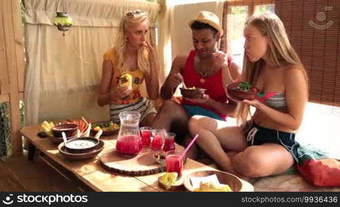 Group of young friends on summer's vacation enjoying a healthy meal in a treehouse at the beach. Smiling people eating on a rustic wooden table and chatting during lunch